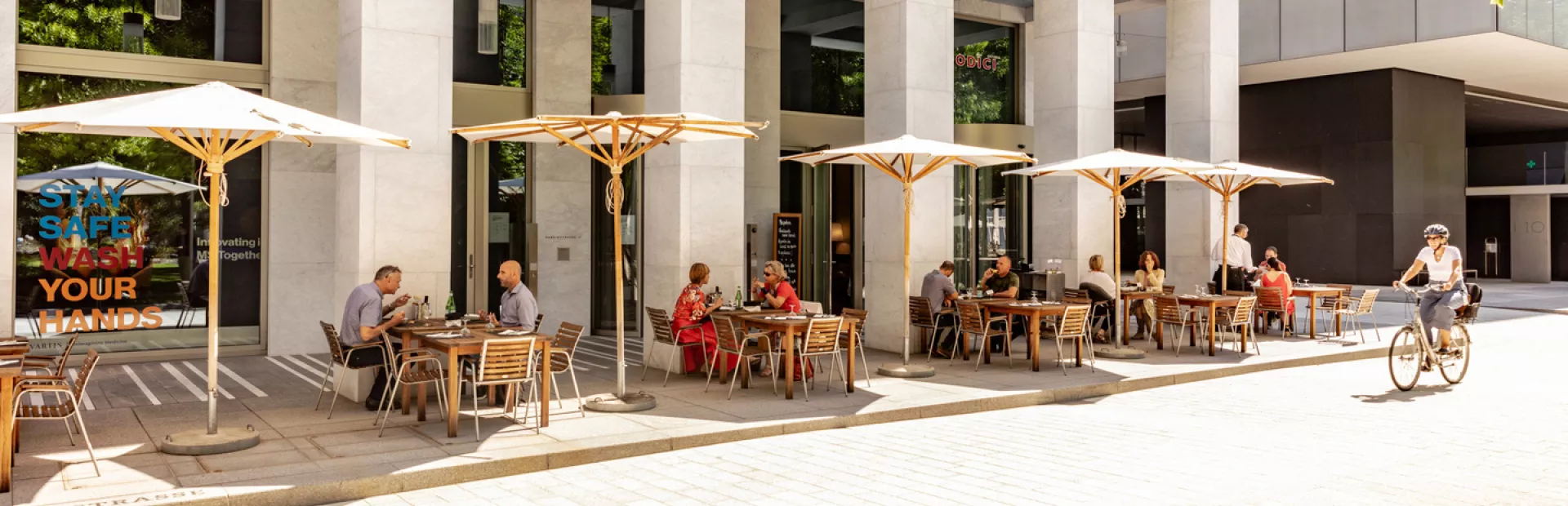 People dining on tables under parasols outside a restaurant on campus on a sunny day