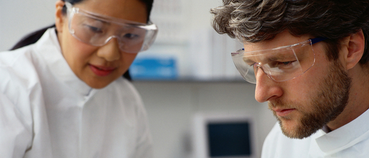 Two scientists wearing lab coats and goggles working together